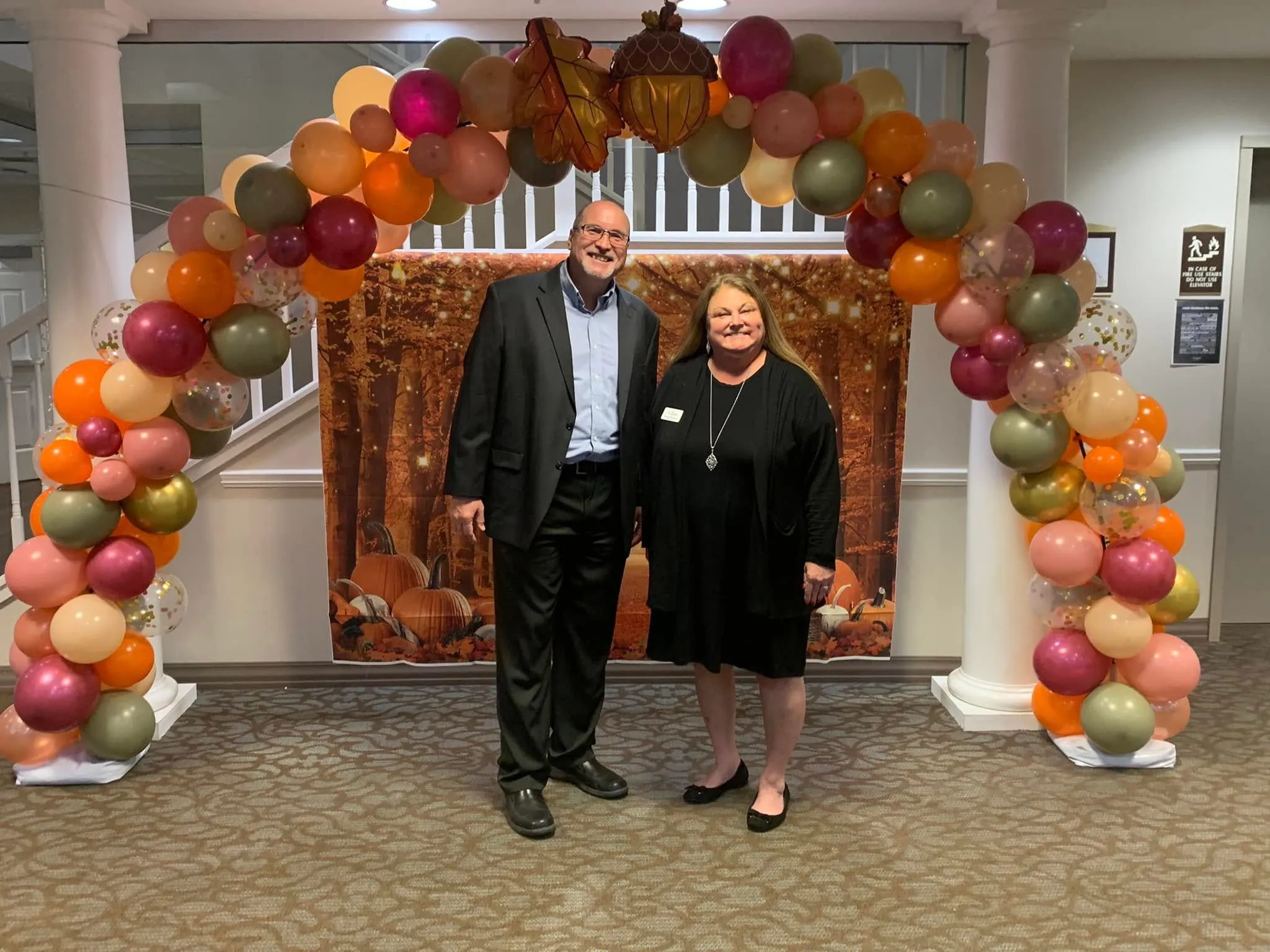 Fall balloon arch in burgundy, sage, peach, orange, blush and gold confetti with leaf and acorn foils over an autumn forest backdrop