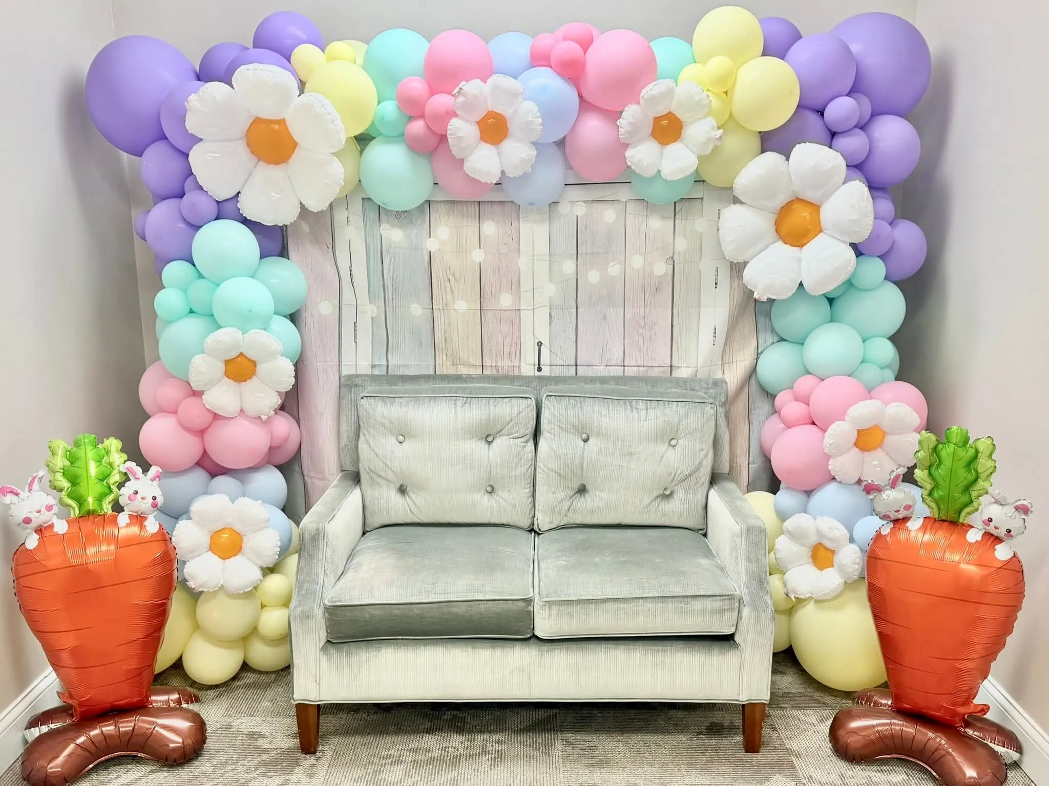 Pastel balloon garland with daisy balloons around a barn backdrop with a loveseat and carrot balloons