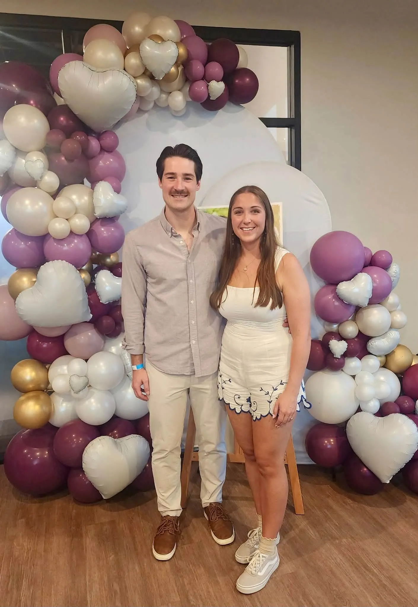 An engaged couple smiling in front of a burgundy, mauve and pearl balloon arch with white heart balloons