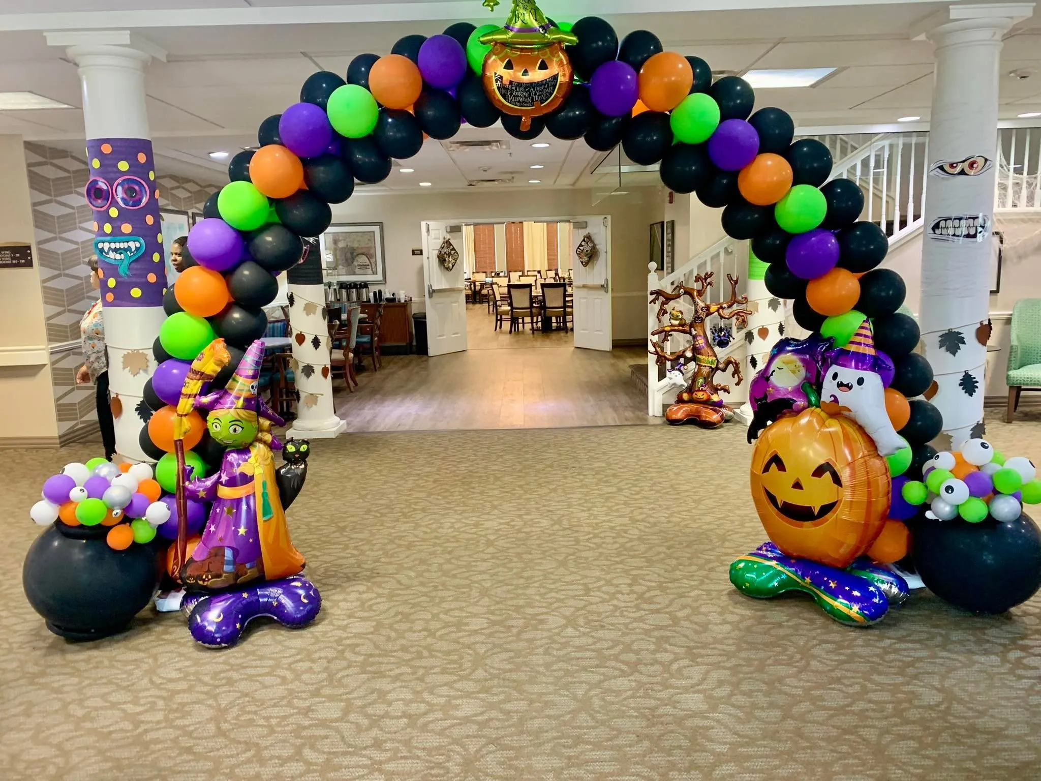 Black, orange, purple and lime balloon arch with a jack-o’-lantern topper, a witch and pumpkin balloon at each side and balloon cauldrons with eyeballs