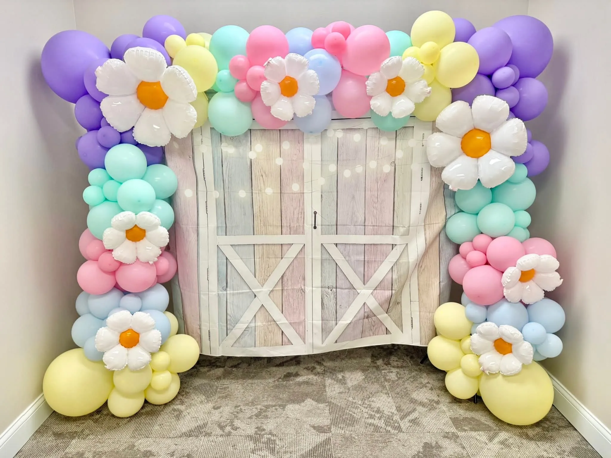 Pastel lavender, mint, pink, blue and yellow balloon garland with white daisy balloons around a pastel barn-door backdrop